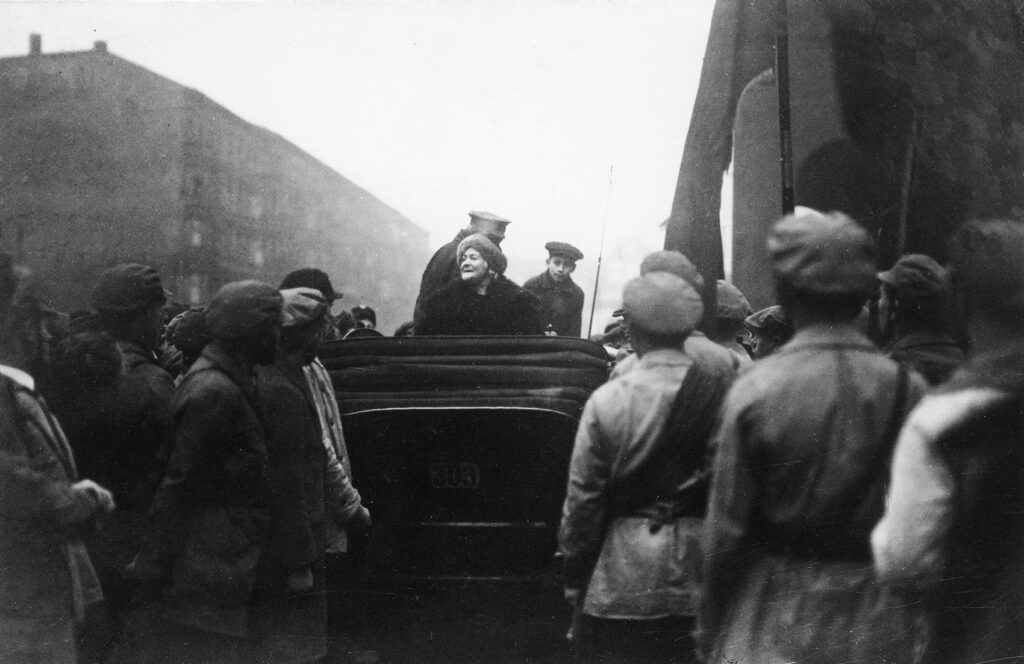 im Vordergrund sieht man Arbeiter und in der Mitte eine Kutsche in der Clara Zetkin sitzt und zu den Arbeitern schaut